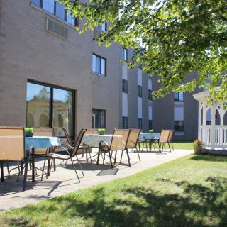 A sunny outdoor patio area with chairs and tables next to a building, featuring a gazebo on a well-maintained lawn.