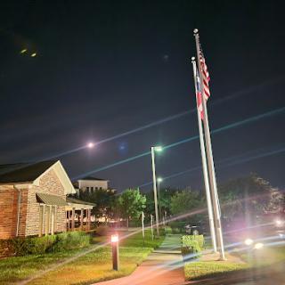An illuminated American flag stands tall against the night sky, surrounded by well-lit pathways and buildings.