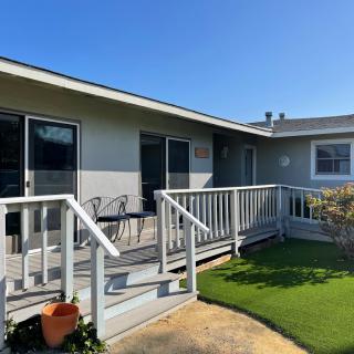 Sunny day view of a house with a wooden deck and railing, featuring outdoor furniture and a garden area.