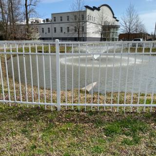 A fenced pond with a central fountain in front of a large building.