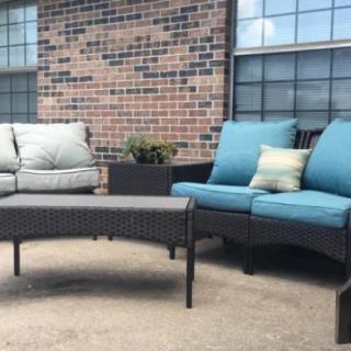 An outdoor patio setup with wicker sofas adorned with blue and gray cushions, a central table, and a backdrop of a brick wall and windows.