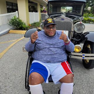 A joyful individual in a wheelchair poses with a vintage car in the background.