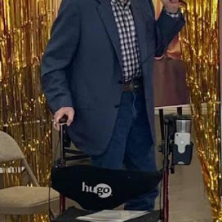 A man in formal attire is standing by a wheelchair, holding a cane and smiling at the camera against a shiny gold backdrop.