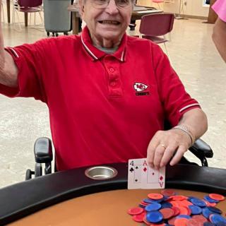 A senior man in a wheelchair proudly holds up his poker hand, surrounded by chips on the table.