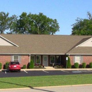 A one-story residential building with brick exterior and parking lot, surrounded by greenery.