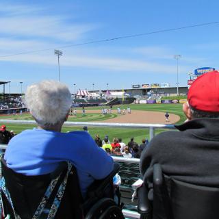 Two people in wheelchairs enjoy a sunny day at a baseball game.