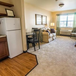 A cozy apartment living room with a mix of carpet and wood flooring, featuring a sofa, small dining table, and a window allowing natural light.