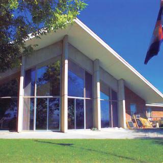 A modern building with large windows and a sloped roof, surrounded by a green lawn and trees.