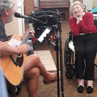 A musician plays the guitar and sings to an elderly woman in a wheelchair, creating a joyful moment.