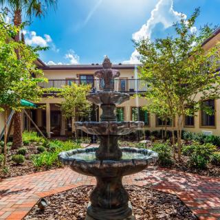 A sunny courtyard with a multi-tiered fountain surrounded by lush greenery and brick pathways.