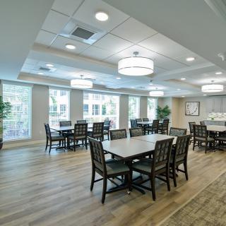 A spacious and well-lit dining area with multiple tables and chairs, featuring large windows and wooden flooring.
