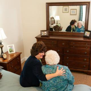 A caregiver comforts an elderly woman in a cozy bedroom setting with a large dresser mirror reflecting their embrace.