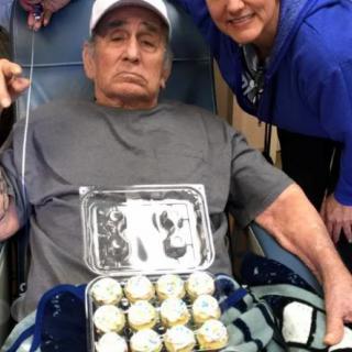 An elderly man in a wheelchair holding cupcakes, joined by a nurse in a hospital setting.