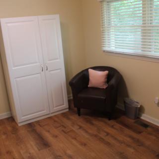 A cozy room featuring a white wardrobe, a black armchair with a pink pillow, and a wooden floor.