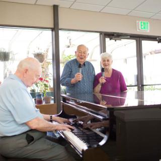 Elderly individuals enjoying music together, with one playing the piano and two others singing.