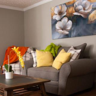 A cozy living room with a grey sofa adorned with colorful cushions and a vibrant throw, complemented by a floral painting on the wall.