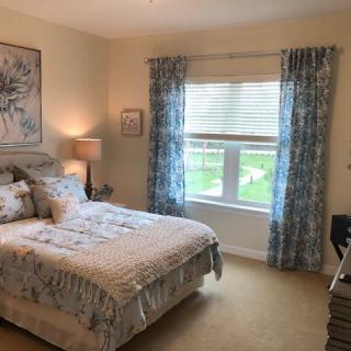 A cozy bedroom with a neatly made bed, curtains framing a large window, and decorative elements like lamps and artwork.