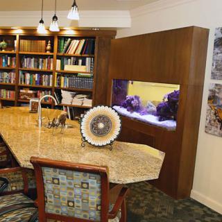 A cozy library setting with a large bookshelf, an aquarium built into the wall, and a central table for reading or study.