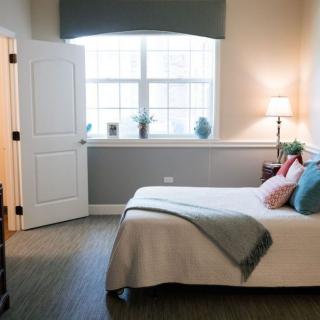 A modern bedroom interior with a neatly made bed, ambient lighting, and natural light streaming through a large window.