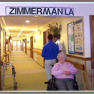 An elderly woman in a wheelchair sits in a hallway of a nursing home, with signage reading 'Zimmerman LA'.