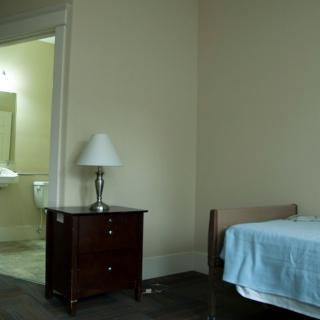 A simple bedroom with a bed, nightstand, and lamp, leading to an adjoining bathroom.