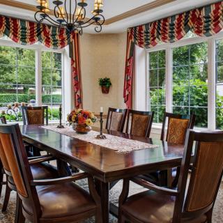 A dining room with a wooden table, surrounded by chairs and adorned with a chandelier, featuring large windows with decorative curtains.