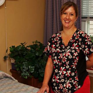 A woman in a healthcare setting, wearing a floral print uniform, stands smiling beside a bed.