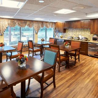 A cozy dining area with wooden tables and chairs, featuring a kitchen space and large windows with decorative curtains.