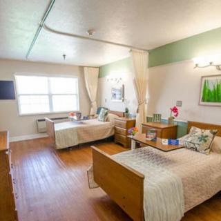A well-lit nursing home room with twin beds and wooden flooring.