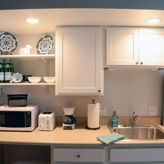 A cozy kitchen corner featuring a microwave, toaster, coffee maker, and sink with white cabinets and shelves displaying dishes.