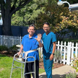 A caregiver assists an elderly individual outdoors, both sharing a moment of joy.