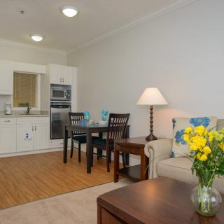 A cozy open-plan living space with a kitchen and dining area, featuring a sofa, table lamp, and a vase of yellow flowers.