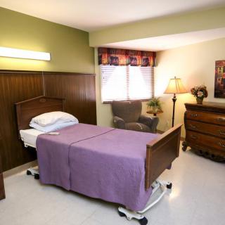 A room featuring a hospital bed with a purple blanket, next to a chair and lamp.
