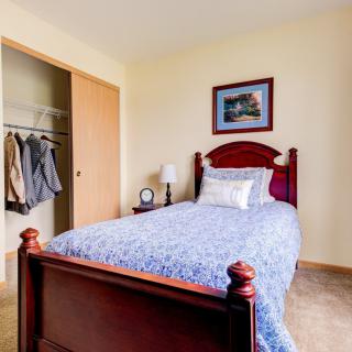 A cozy bedroom featuring a single bed with a blue patterned cover, a bedside lamp, an open closet with hanging clothes, and artwork on the wall.