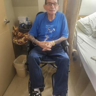 A man in a wheelchair wearing a blue shirt and displaying tattoos, seated in a cozy indoor environment.