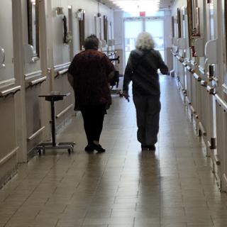 Two elderly individuals walking together in a hallway of a care facility.