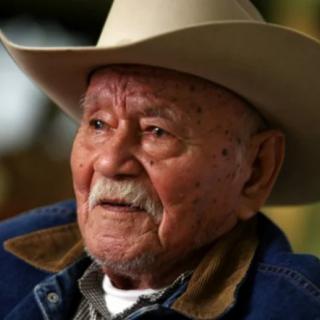 An elderly man wearing a cowboy hat looks thoughtfully into the distance.