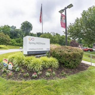 A landscaped area with a sign surrounded by bushes and plants, displaying an American flag and a lamp post in the background.
