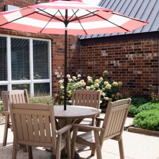 A cozy patio setup with a round table and chairs under a red and white striped umbrella, surrounded by a garden with brick walls.