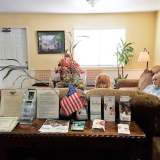 A waiting room with people sitting on sofas, surrounded by brochures, plants, and a flag.