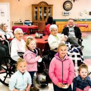 A group of children and seniors gather together indoors, sharing smiles and warmth.