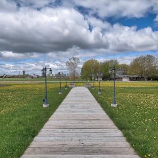 A wooden pathway flanked by lampposts stretches through a green field under a partly cloudy sky.