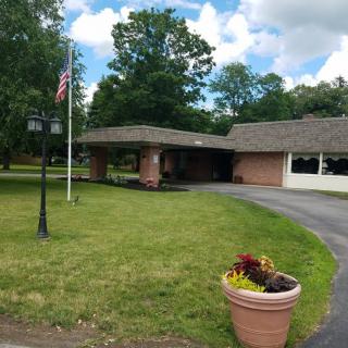 A suburban house with a covered driveway, surrounded by trees and a manicured lawn, featuring a flagpole and decorative flower pot.