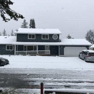 A suburban house covered in snow on a winter day, with parked cars and trees surrounding the area.