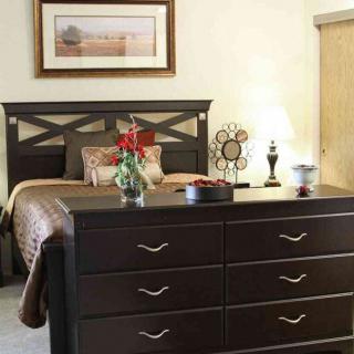 A cozy bedroom featuring a bed with a dark headboard, a large dresser, and decorative items, including a lamp and flowers.