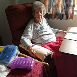 An elderly woman sitting comfortably in her armchair, surrounded by personal items in a sunlit room.