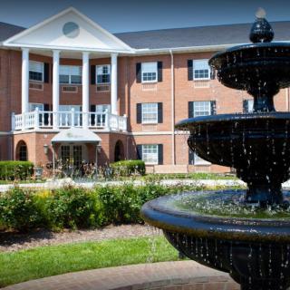 A traditional brick building with white columns stands behind a decorative fountain amidst a landscaped garden.