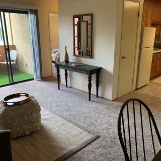 A cozy living room with carpeted flooring, featuring a table and mirror, adjacent to a kitchen and balcony.