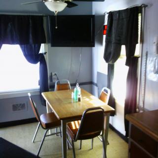 A small dining room with a wooden table and four chairs, featuring two bottles on the table and dark curtains on the windows.