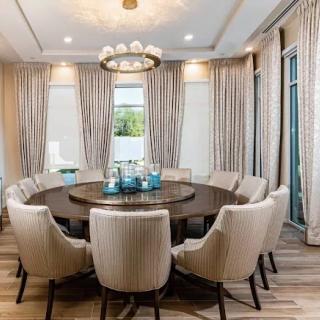 A modern dining room featuring a round table surrounded by elegant chairs, with a chandelier above and soft curtains framing the windows.